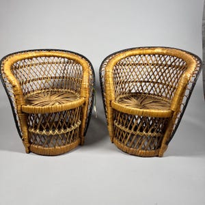 Op de afbeelding: Twee vintage rotan stoelen met een geweven ontwerp. De stoelen hebben een afgeronde rug en zitting, met een zwarte rand rond de randen. Het rotan is een natuurlijke bruine kleur en de stoelen staan tegen een grijze achtergrond.