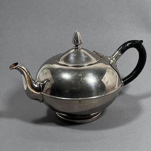 Könnte beinhalten: Eine silberne Teekanne mit schwarzem Griff und dekorativem Knauf auf dem Deckel. Die Teekanne hat einen runden Körper und einen gebogenen Ausguss. Die polierte Oberfläche reflektiert die Umgebung.