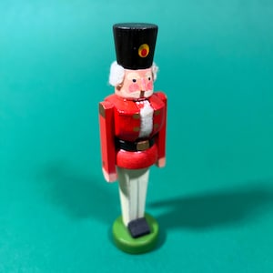 Peut inclure: Figurine de casse-noisette en bois, représentant un soldat classique. Il arbore une veste rouge, un chapeau noir avec un accent doré et un pantalon blanc. La figurine repose sur une base verte et a un visage peint détaillé.