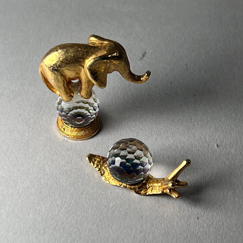 May include: Two decorative figurines: a gold-colored elephant atop a faceted crystal ball and a gold-colored snail with a faceted crystal ball shell. The elephant figurine has detailed features, and the snail has extended antennae.