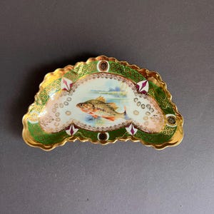Könnte beinhalten: Eine halbmondförmige Porzellanschale mit einer handgemalten Illustration eines Fisches in einem See in der Mitte. Der Rand ist mit einem tiefgrünen Muster, goldenen Filigranakzenten und kleinen magentafarbenen Rauten verziert.