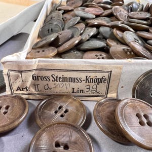May include: A collection of brown buttons with four holes, some with a textured surface, are displayed in a cardboard box. The box has a label with German text. The buttons vary in shades of brown and gray.