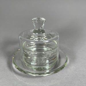 May include: A clear glass butter dish with a lid and a round base. The dish has a cylindrical shape with a small handle on top of the lid. The base is a flat, circular plate. The glass is transparent, allowing a view of the contents.