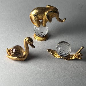 May include: Three decorative figurines: a gold elephant atop a crystal ball, a gold swan, and a gold snail with a crystal ball shell. The figurines are set against a neutral background, showcasing their intricate details and craftsmanship.