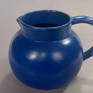 May include: A solid blue ceramic jug with a rounded body and a handle. The jug is glazed and has a smooth finish.