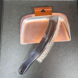 Könnte beinhalten: Ein Vintage-Staubwisch- und Bürstenset. Die Kehrschaufel ist kupferfarben mit einem Holzgriff, und die Bürste hat einen schwarzen Griff und weiße Borsten. Das Set ist zum Aufkehren von Staub und Schmutz bestimmt.