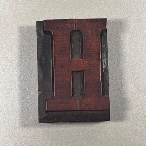 May include: A vintage wooden letterpress printing block featuring the capital letter "H". The letter is a dark reddish-brown color, set within a black frame. The block shows signs of wear, indicating its use in printing.