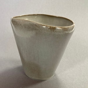 May include: A white ceramic vase with a slightly irregular, rounded shape. The vase has a light brown glaze and a smooth, glossy finish.
