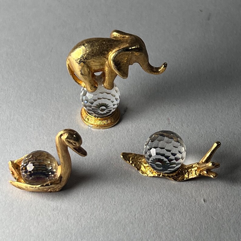 May include: Three decorative figurines: a gold elephant atop a crystal ball, a gold swan with a crystal body, and a gold snail with a crystal shell. The figurines are set against a neutral background.