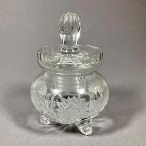 Könnte beinhalten: Eine klare Glasdose mit Deckel und drei Beinen. Die Dose hat ein geschliffenes Glasdesign mit einem Blumenmuster.