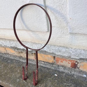 Op de afbeelding: Een roestbruine metalen wandhanger met een cirkelvormig ontwerp en twee haken. Het metaal vertoont tekenen van veroudering. Het ontwerp heeft een grote ring aan de bovenkant en twee verticale staven die eindigen in haken.