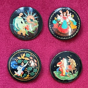 Könnte beinhalten: Vier runde, schwarz lackierte Schachteln mit farbenfrohen, detaillierten Illustrationen. Jede Schachtel zeigt eine andere Szene mit Figuren in traditioneller Kleidung. Die Schachteln sind auf einer rosa Oberfläche angeordnet.