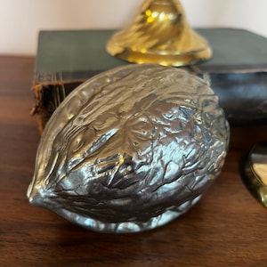Puede incluir: Un cascanueces de metal plateado con forma de nuez. El cascanueces tiene una superficie texturizada y está parcialmente enfocado. Un objeto de color dorado es visible en el fondo.