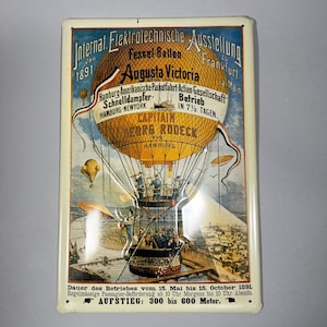 May include: Vintage metal sign featuring a hot air balloon with passengers, titled "Augusta Victoria." The sign includes German text and dates from 1891, with a cityscape in the background. The sign is rectangular with a cream-colored border.