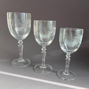 Könnte beinhalten: Drei klare Weingläser mit geätzten floralen Mustern. Die Gläser haben eine abgerundete Schale und einen schlanken Stiel. Die Gläser sind in verschiedenen Größen erhältlich. Sie werden auf einer grauen Oberfläche präsentiert, was ihr zartes Design hervorhebt.
