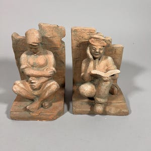 May include: Set of two terracotta bookends. Each bookend features a seated figure, one with arms crossed and the other reading a book. The figures are set against a brick-like backdrop. The bookends are a light brown color.