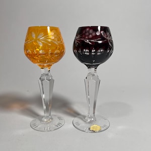 May include: Two crystal wine glasses. One is amber, the other is deep red. Both have etched floral and grape designs on the bowls. The stems are clear, faceted, and the bases are round.