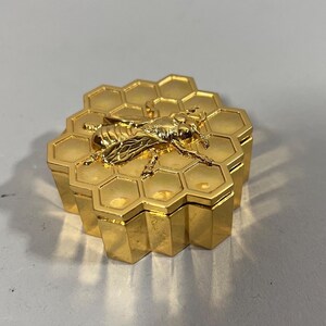 Può includere: Una scatola esagonale color oro con un'ape dettagliata sopra. La scatola è progettata per assomigliare a un favo. L'ape è finemente dettagliata, con ali e segmenti del corpo visibili. La scatola è realizzata in un materiale metallico lucido.
