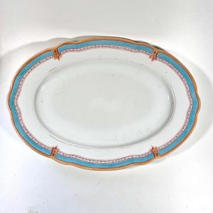 Könnte beinhalten: Ovale weiße Servierplatte mit gewelltem Rand. Der Rand hat ein türkisfarbenes Band mit roten und goldenen Akzenten. Ein klassisches Stück zum Servieren von Speisen bei einem Abendessen oder besonderen Anlässen.
