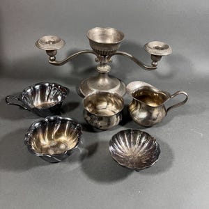 Könnte beinhalten: Eine Auswahl an antiken Servierteilen aus Silber, darunter ein zweiarmiger Leuchter, ein Sahnekännchen und mehrere kleine Schalen. Die Teile haben eine angelaufene Oberfläche, und die Schalen haben dekorative Ränder.