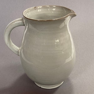Può includere: Un'anfora in ceramica bianca con corpo arrotondato e manico. L'anfora ha una superficie leggermente strutturata e un bordo marrone.