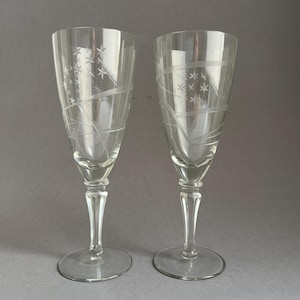 May include: Two clear champagne flutes with etched star and swirl designs. The flutes have a tall, slender shape with a round base and a detailed stem. The etched design adds a decorative element to the glassware.