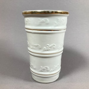 Puede incluir: Jarrón de cerámica blanca con borde dorado y diseño floral en relieve. El jarrón está dividido en tres secciones por bandas doradas.