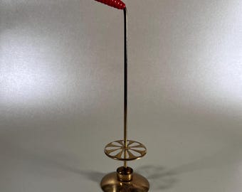 May include: A vintage brass candle holder with a red handle. The candle holder has a circular base, a central stem, and a decorative floral-shaped element. The red handle is curved and has a textured surface.