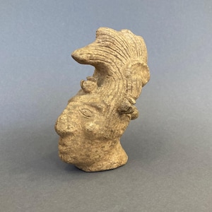 Könnte beinhalten: Ein hellbrauner, geformter Kopf mit detaillierten Gesichtszügen und einem stilisierten Kopfschmuck. Die Skulptur hat ein verwittertes Aussehen, das auf Alter oder eine rustikale Ästhetik hindeutet. Der Kopf ist im Profil positioniert und zeigt das komplizierte Design.