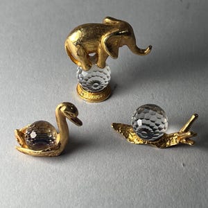May include: Three small decorative figurines: a gold elephant atop a crystal ball, a gold swan with a crystal body, and a gold snail with a crystal shell. The figurines are on a light gray surface.