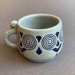 Puede incluir: Una taza pequeña de cerámica gris con un diseño en espiral azul y blanco. La taza tiene un asa con una bisagra de metal.