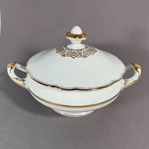 Peut inclure: Soupière en porcelaine blanche avec garniture et poignées dorées. Le couvercle présente un motif floral doré et un bouton décoratif. La soupière a un bord festonné et est conçue pour servir de la soupe.