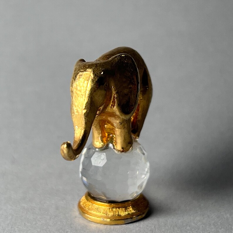 May include: A gold-colored elephant figurine balancing on a clear, faceted crystal ball. The elephant has detailed features and stands on a gold-colored base. The figurine is a decorative item.