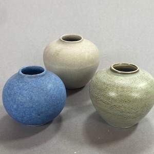 May include: Three small ceramic vases in different colours. The vase on the left is blue, the vase in the centre is beige, and the vase on the right is green.