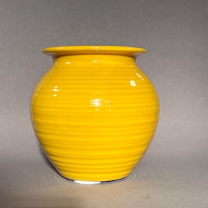 Könnte beinhalten: Eine leuchtend gelbe Keramikvase mit rundem Korpus und leicht ausgestelltem Rand. Die Vase hat horizontale geriffelte Details um ihre Außenseite. Der Sockel ist heller gelb. Ein dekorativer Gegenstand.