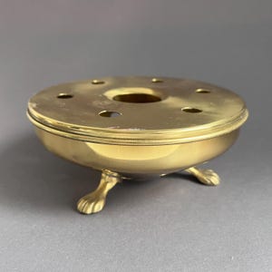 Puede incluir: Un calentador de té de latón con una parte superior redonda con múltiples agujeros y una abertura central. El calentador descansa sobre tres patas decorativas con forma de garra. El diseño es de color dorado.