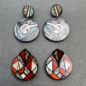 Puede incluir: Cuatro pares de pendientes. Dos pares son circulares con bordes negros y presentan diseños geométricos en tonos grises, blancos y naranjas. Los otros dos pares tienen una base circular con un diseño geométrico en rojo, blanco y negro.