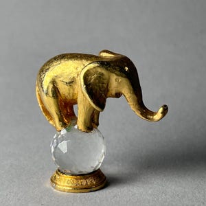 May include: A gold-colored elephant figurine balancing on a clear, faceted crystal ball. The elephant has detailed features and is standing on a round, gold-colored base. The background is a solid gray.