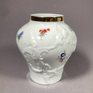 Puede incluir: Jarrón de cerámica blanca con borde dorado y volutas en relieve. El jarrón está decorado con diseños florales coloridos en rojo, azul y morado. Una pieza decorativa para la decoración del hogar.