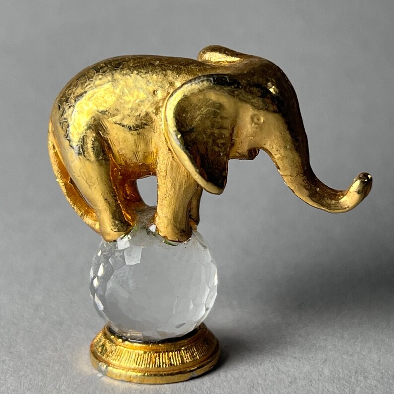 May include: A gold-colored elephant figurine standing on a clear crystal ball. The elephant has detailed features and is perched on a faceted sphere. The base of the sphere is gold, creating a decorative accent.