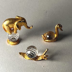 May include: Three small, decorative figurines: a gold elephant atop a crystal ball, a gold swan with a crystal body, and a gold snail with a crystal shell. The figurines are set against a neutral background.
