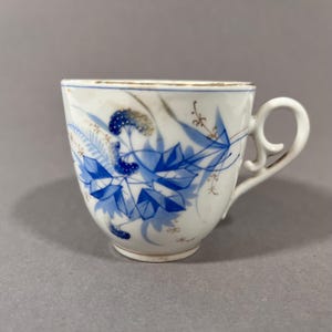 May include: White teacup with a blue floral design. The cup features a curved handle and a gold rim. The blue design includes leaves and other floral elements. The cup is set against a gray background.