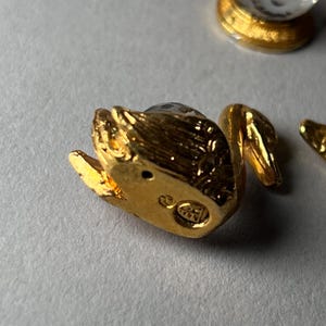 May include: A small, gold-colored swan figurine with a clear crystal head. The swan has a detailed, textured surface and is positioned on a light gray surface. The swan's beak and wings are visible.