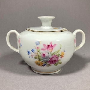 Puede incluir: Azucarero de porcelana blanca con un diseño floral. El tazón tiene una tapa y dos asas. Las flores son rosas, azules, amarillas y verdes.