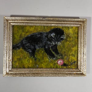May include: Oil painting of a black kitten playing with a pink ball in a field of green grass. The artwork is in a gold-coloured ornate frame. The kitten is the focal point of the artwork.