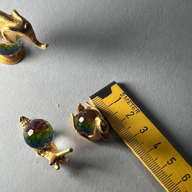 May include: Gold-toned snail-shaped earrings with a faceted, rainbow-colored glass bead. A yellow ruler is next to the earrings. The earrings are on a gray surface. The image also shows a gold-toned elephant figurine.