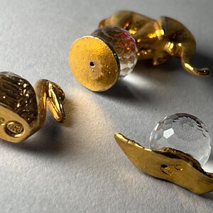 May include: A collection of gold-colored decorative objects, including a small elephant figurine, a bottle stopper with a faceted crystal ball, and a leaf-shaped piece holding a clear crystal sphere. The objects are arranged on a light gray surface.