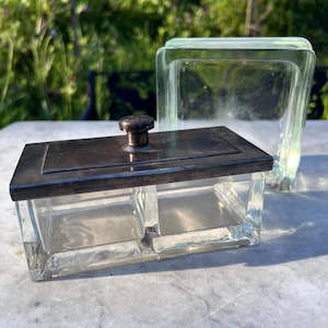 Könnte beinhalten: Eine rechteckige Kiste aus klarem Glas mit einem silberfarbenen Deckel und einem kleinen, runden Knopf. Die Kiste ist in zwei Fächer unterteilt. Dahinter befindet sich ein größerer, quadratischer Behälter aus klarem Glas. Beide stehen auf einer hellen Oberfläche.