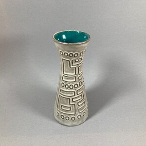 Könnte beinhalten: Eine weiße Keramikvase mit einem blaugrünen Überzug im Inneren. Die Vase hat ein geprägtes geometrisches Muster auf der Außenseite.