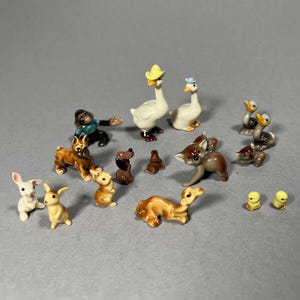 May include: A collection of small ceramic animal figurines, including geese, ducks, rabbits, dogs, and deer. The figurines are painted in various colours and feature detailed designs. The animals are arranged on a grey surface.